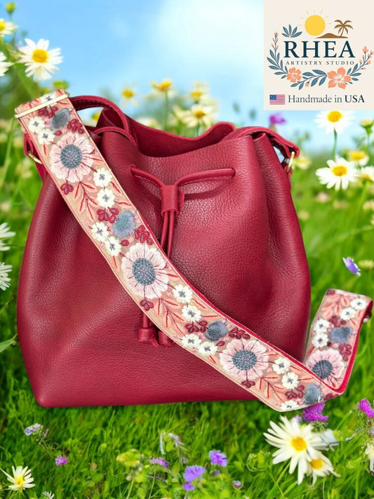 Rhea Artistry Studio 2-inch crossbody strap with embroidered floral lace in pink, white, and blue on a tan background, attached to a burgundy bucket bag.