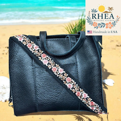 Rhea Artistry Studio black leather handbag with a 1.5-inch adjustable crossbody strap featuring blush pink and ivory floral embroidery on black webbing.