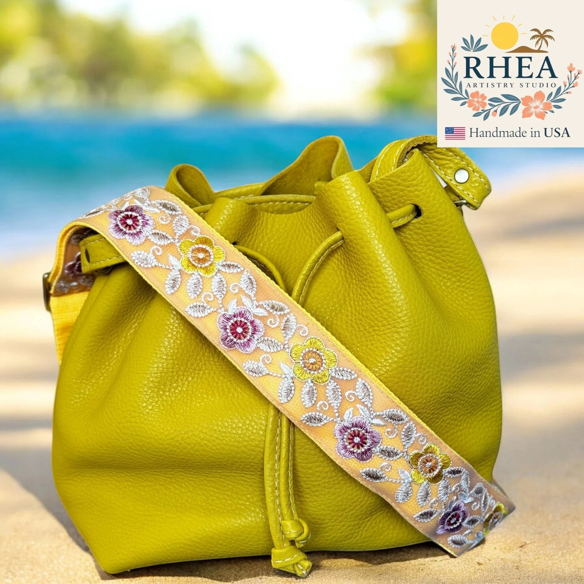 Rhea Artistry Studio 2-inch crossbody strap with embroidered colorful florals on a light yellow background, attached to a lime green leather bucket bag on a beach.