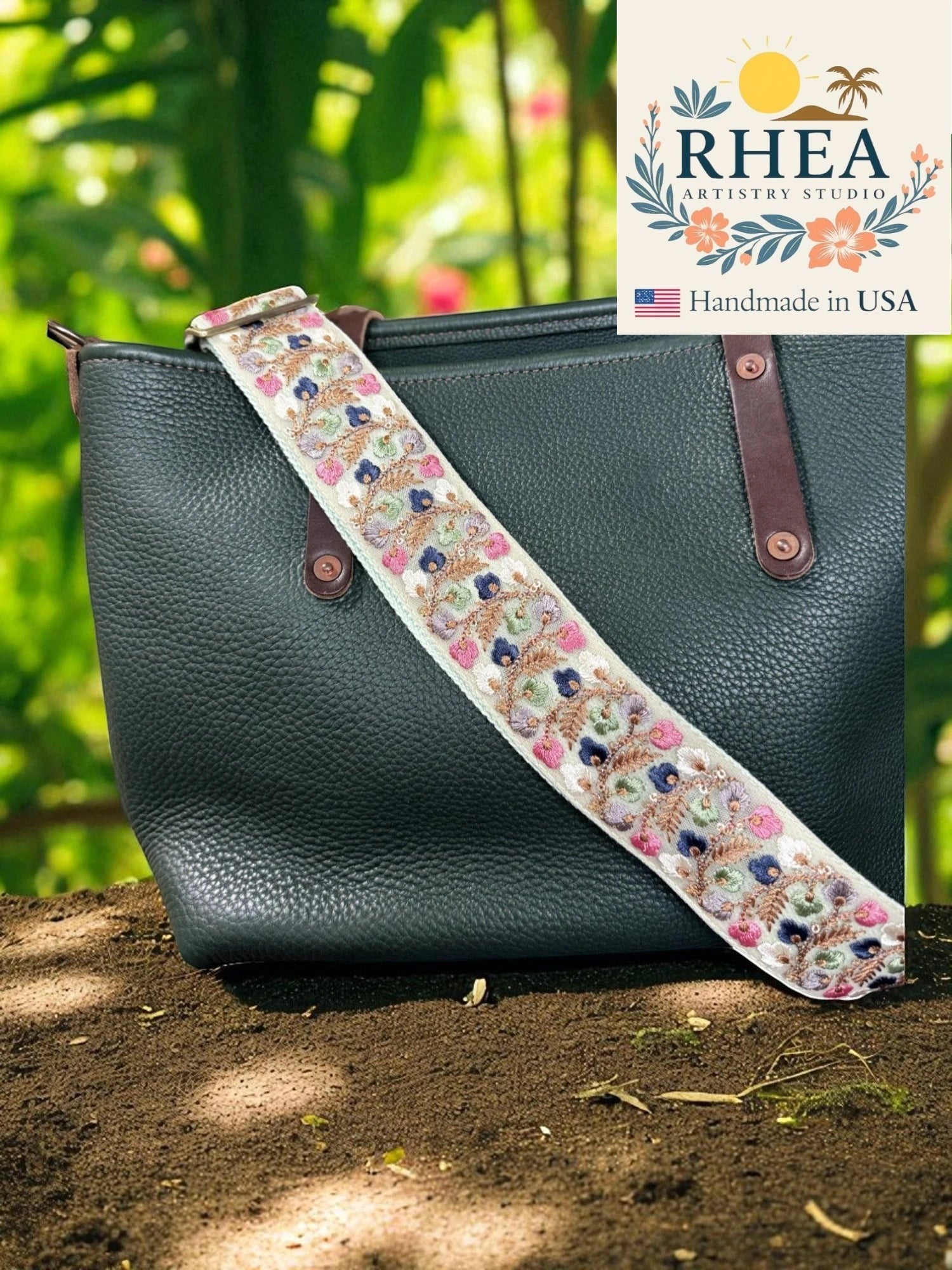 Rhea Artistry Studio 2-inch wide crossbody strap with embroidered floral lace in pink, blue, and green on a cream background, attached to a black leather tote bag.