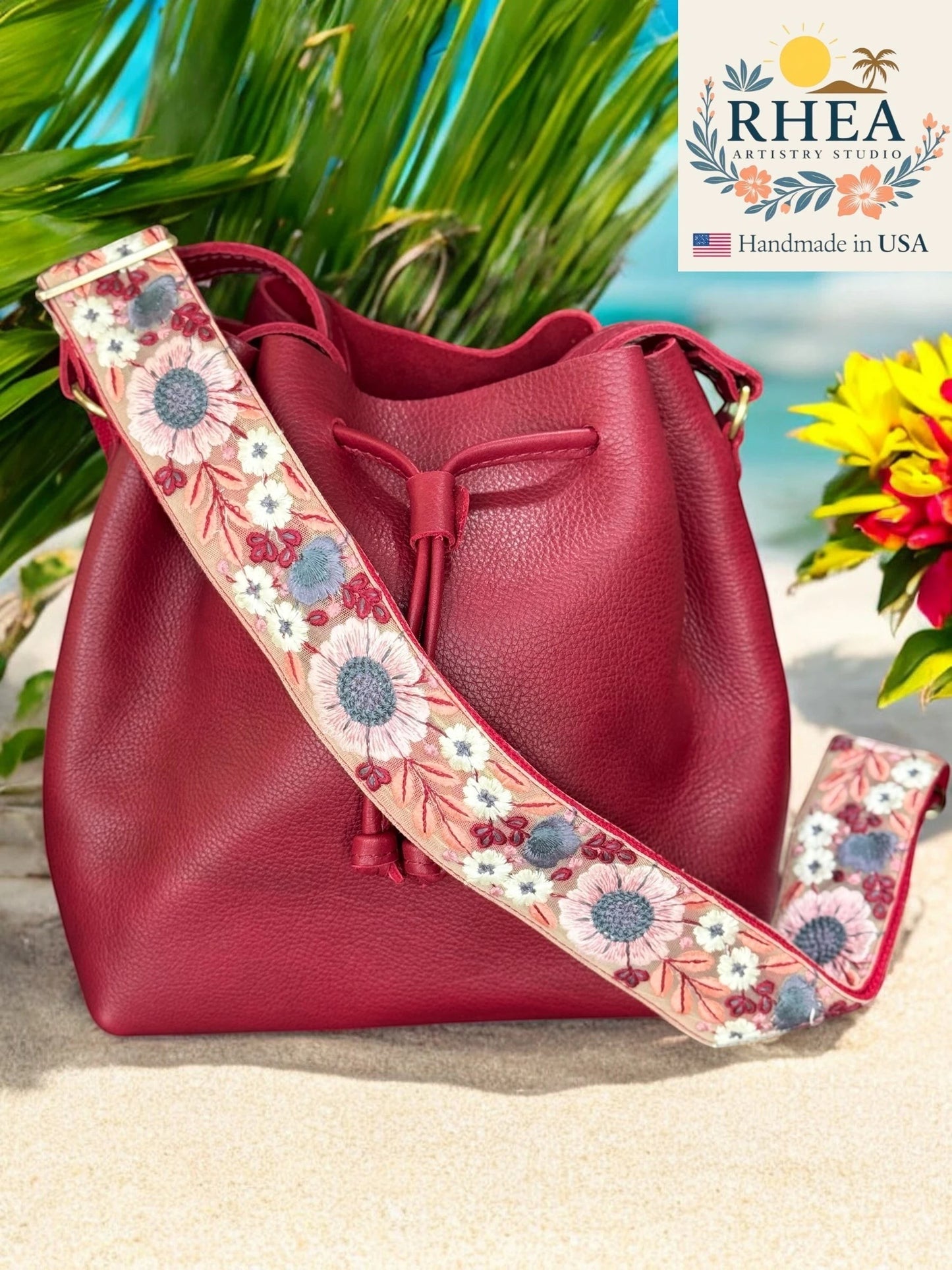Rhea Artistry Studio 2-inch wide crossbody strap with embroidered floral lace in pink, blue, and white on a tan background, attached to a red leather bucket bag.