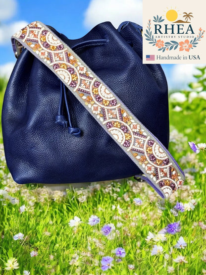 Rhea Artistry Studio 2-inch wide crossbody strap with embroidered floral lace in purple, gold, and cream on a grey background, attached to a navy blue bag.