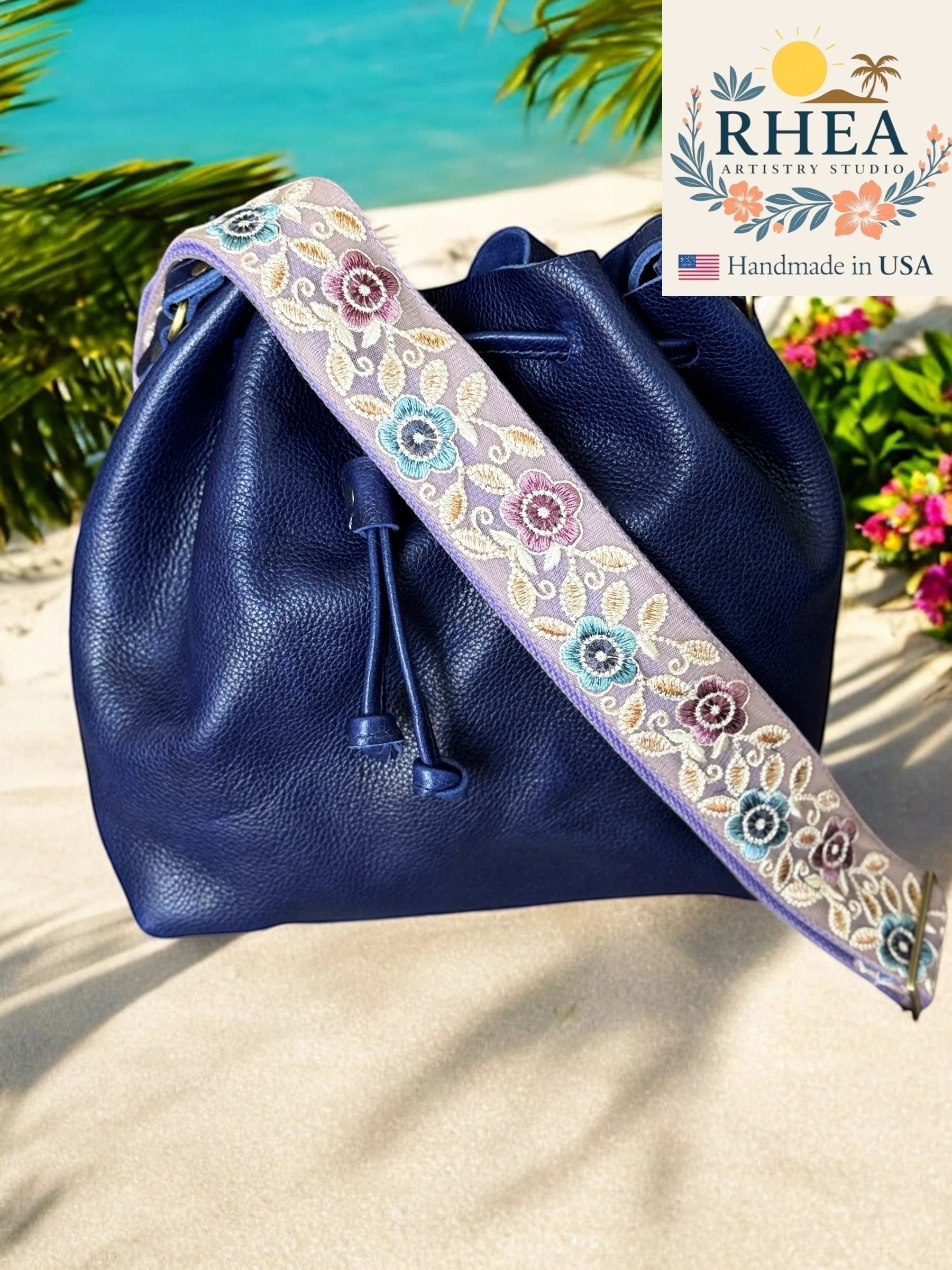 Rhea Artistry Studio 2-inch crossbody strap with embroidered pastel florals on a light purple background, attached to a navy blue leather bucket bag.
