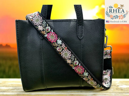 Rhea Artistry Studio 1.5-inch crossbody strap with embroidered pinwheel flowers in pink, white, and green on black webbing. Adjustable length with brass hooks.