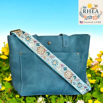 Rhea Artistry Studio light blue crossbody strap with embroidered pinwheel flowers in tan, white, and purple, 1.5-inch wide, adjustable 30-54 inches.
