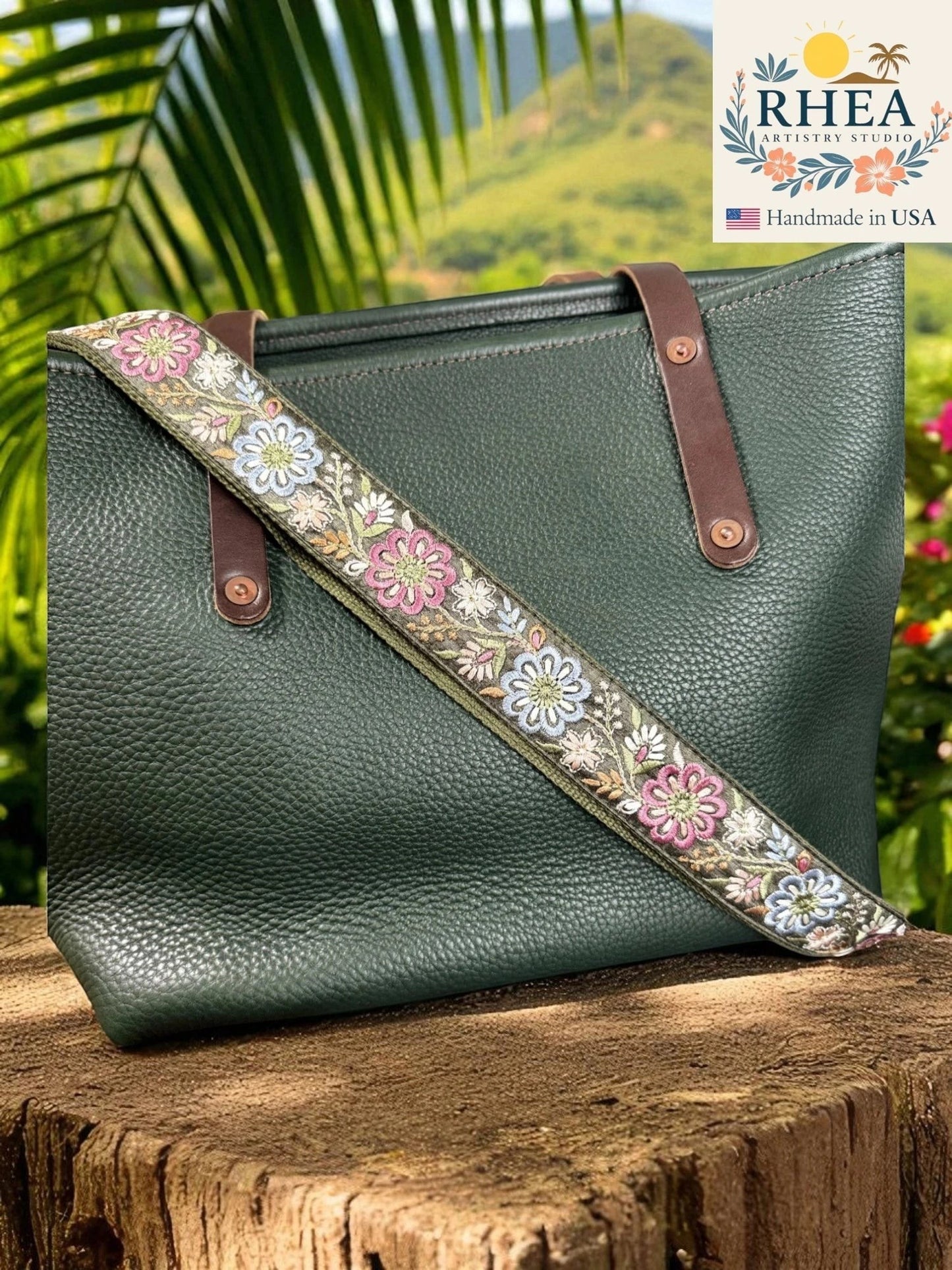 Rhea Artistry Studio 1.5-inch crossbody strap with embroidered pinwheel flowers in pink, blue, and white on a green background, with brushed brass hardware.