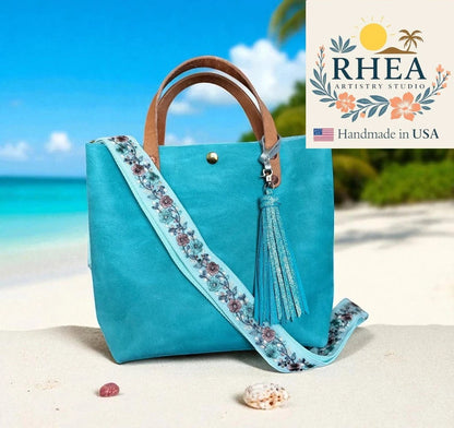 Rhea Artistry Studio adjustable 1.5-inch crossbody strap with blush bloom embroidered floral lace on light blue webbing, brushed brass hardware, and tassel.