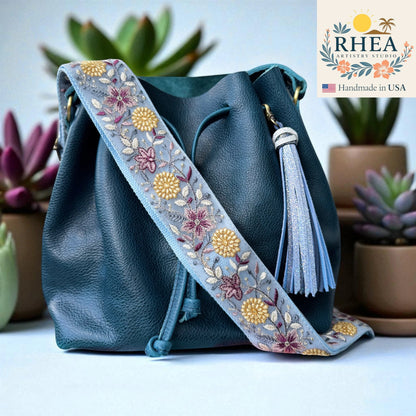 Rhea Artistry Studio 2-inch wide crossbody strap with light blue lace embroidery of floral and leaf motifs in gold, purple, and pink. Adjustable strap with brushed brass hardware.