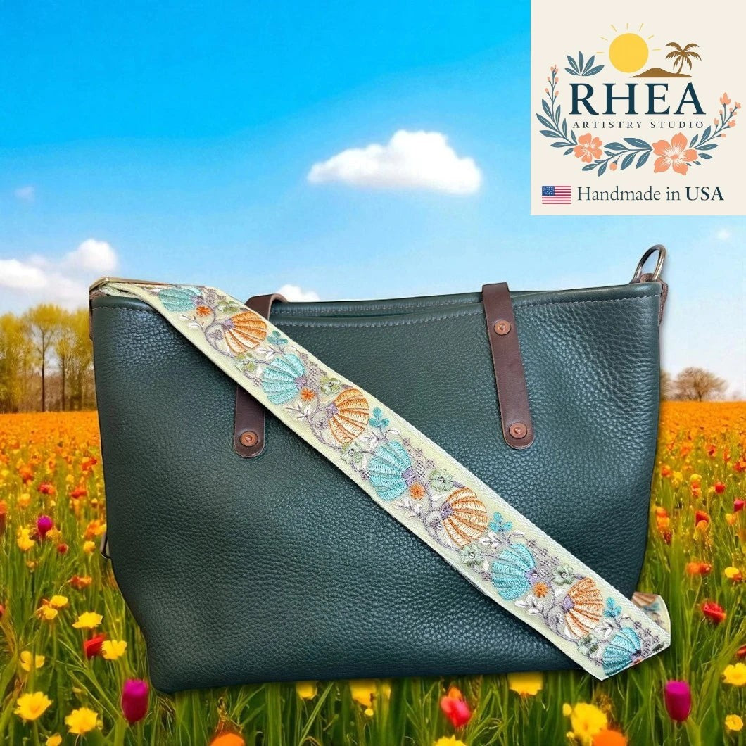 Rhea Artistry Studio 2-inch wide crossbody strap with embroidered shells in blue, orange, and purple on a cream background, attached to a dark green pebbled leather tote bag.