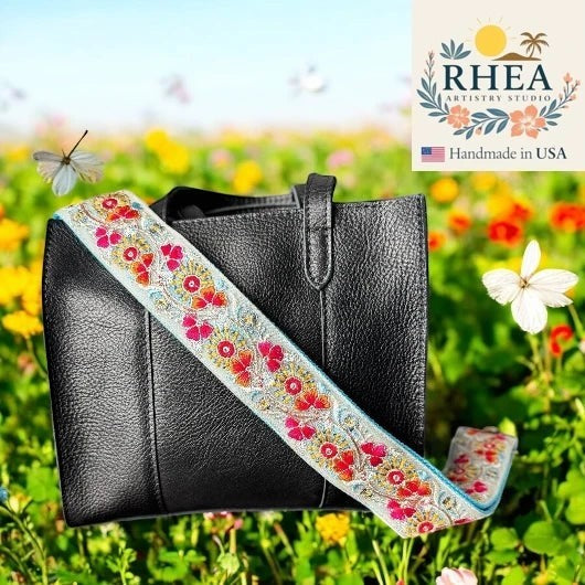 Rhea Artistry Studio 2-inch wide crossbody strap with embroidered Trillium flowers in pink and orange on a light blue background, with brushed brass hardware.