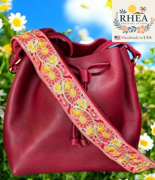Rhea Artistry Studio red leather bucket bag with a 2-inch wide adjustable crossbody strap featuring embroidered trillium flowers in yellow and pink.