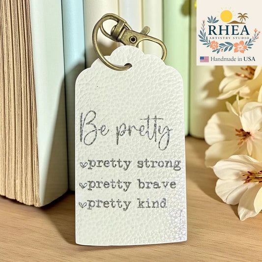 Rhea Artistry Studio white Saffiano leather tag engraved with "Be pretty, pretty strong, pretty brave, pretty kind" affirmations. Includes antique brass clip.