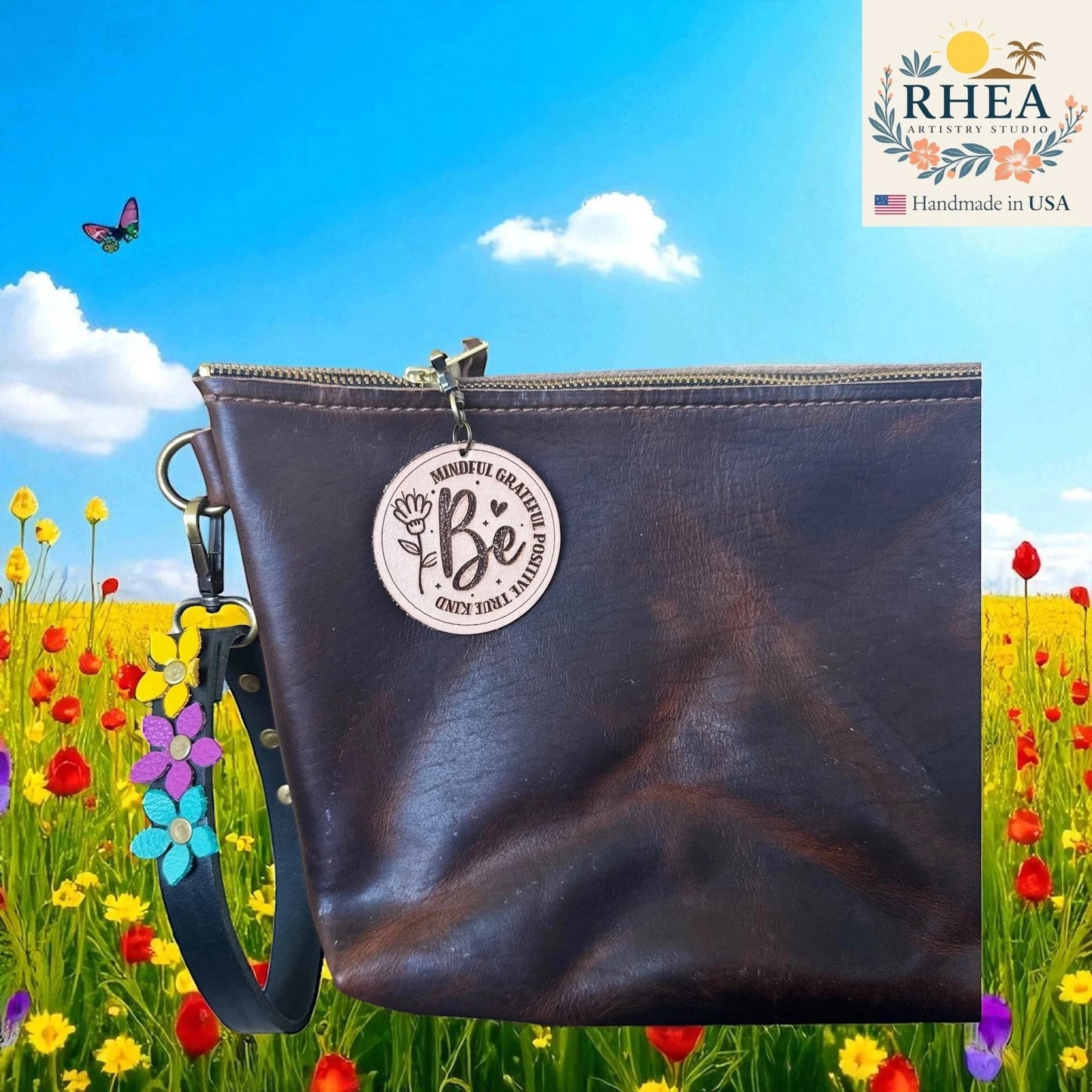 Rhea Artistry Studio round leather tag engraved with 'Be Mindful Grateful Positive True Kind' and a flower design, attached to a dark brown leather bag.