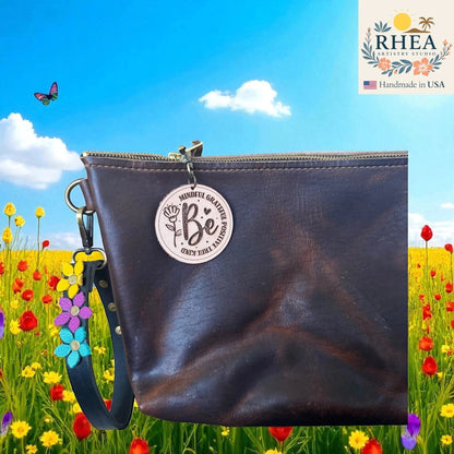 Rhea Artistry Studio round leather tag engraved with 'Be Mindful Grateful Positive True Kind' and a flower design, attached to a dark brown leather bag.