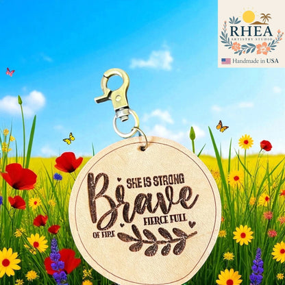 Rhea Artistry Studio natural leather tag engraved with 'She is Strong Brave Fierce Full of Fire' quote, attached to a metal clasp, set against a backdrop of a sunny meadow with flowers and butterflies.