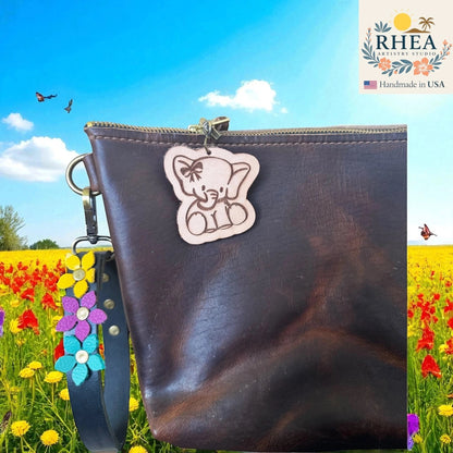 Rhea Artistry Studio natural leather tag engraved with a cute elephant wearing a bow, attached to a dark brown leather bag.