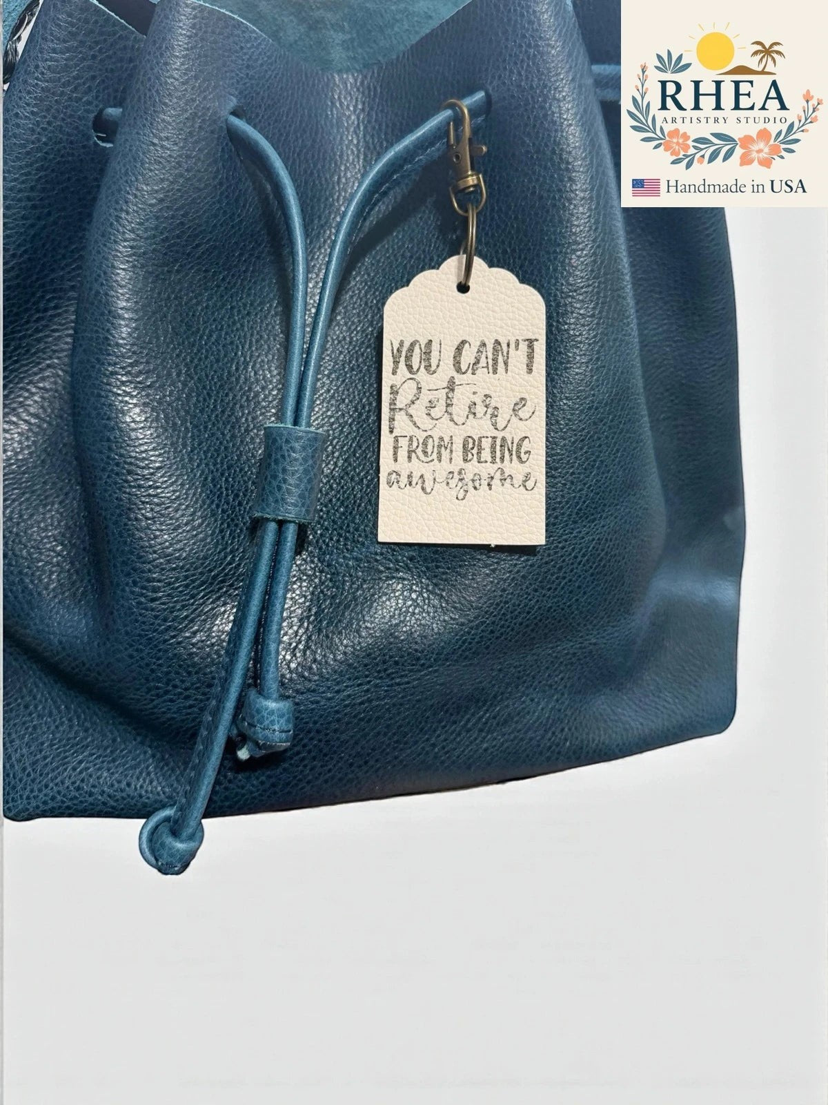 Rhea Artistry Studio cream Saffiano leather tag engraved with "YOU CAN'T Retire FROM BEING awesome" hangs from a teal leather drawstring bag.