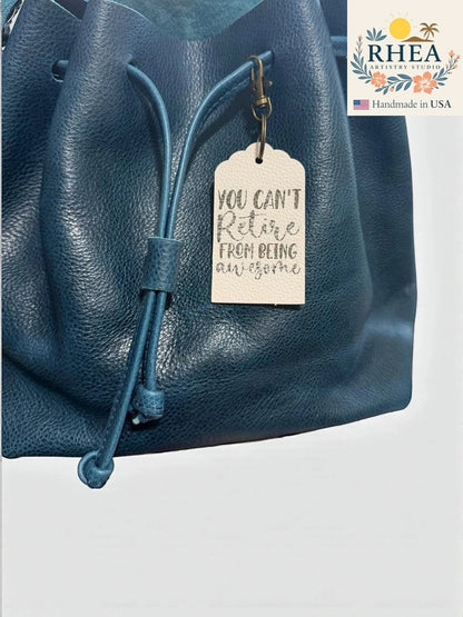 Rhea Artistry Studio cream Saffiano leather tag engraved with "YOU CAN'T Retire FROM BEING awesome" hangs from a teal leather drawstring bag.