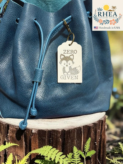 Rhea Artistry Studio cream Saffiano leather tag with engraved fox and "Zero Given" text, clipped to a teal blue leather drawstring bag.
