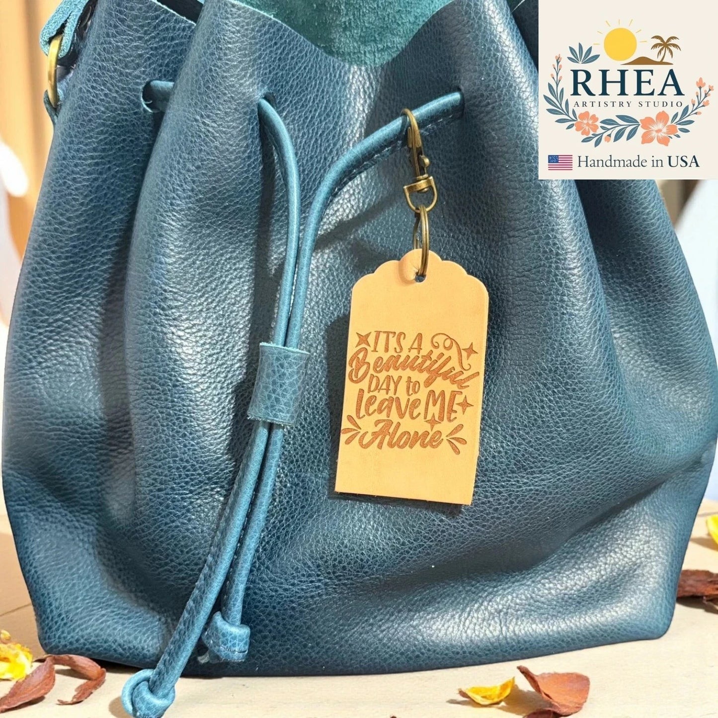 Rhea Artistry Studio natural leather tag engraved with 'It's a beautiful day to leave me alone' quote, attached to a teal blue leather drawstring bag.