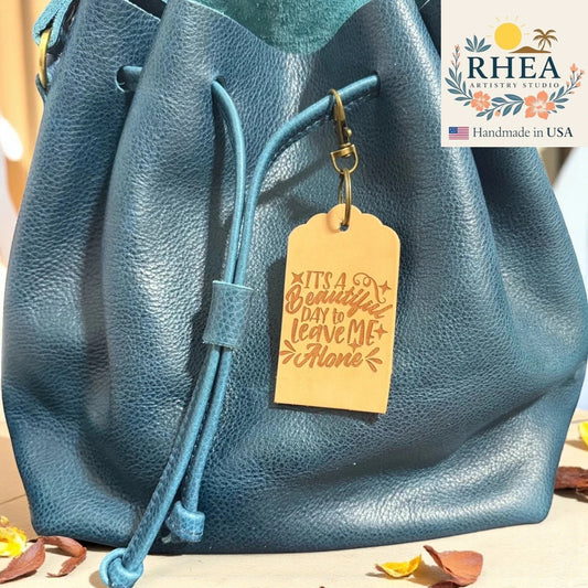 Rhea Artistry Studio natural leather tag engraved with 'It's a beautiful day to leave me alone' quote, attached to a teal blue leather drawstring bag.