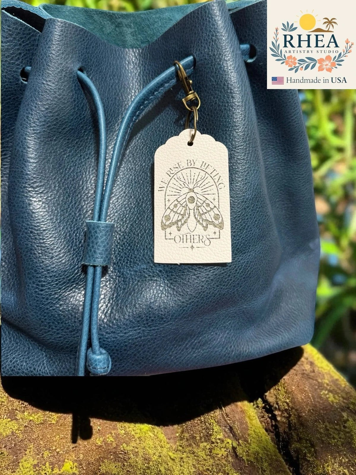 Rhea Artistry Studio navy blue Saffiano leather drawstring bag with a white engraved leather tag featuring a moth and the text "We rise by lifting others."