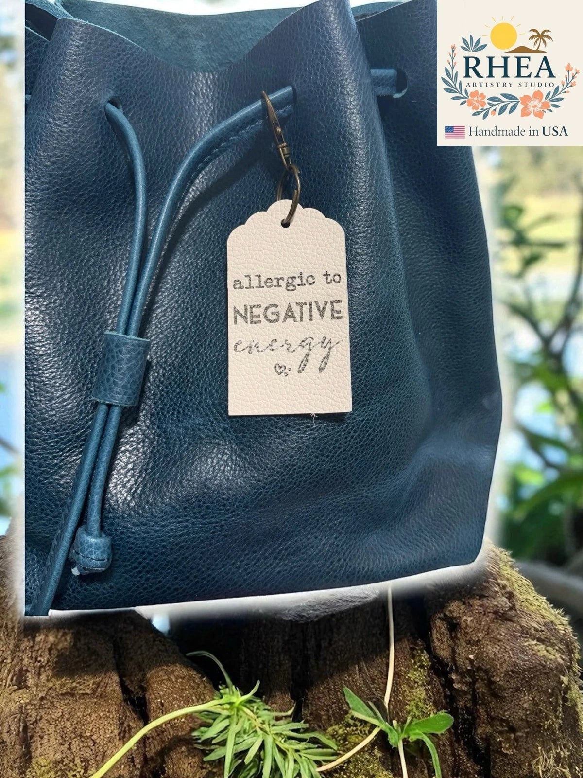 Rhea Artistry Studio cream Saffiano leather tag engraved with 'allergic to negative energy' attached to a teal blue leather drawstring bag.