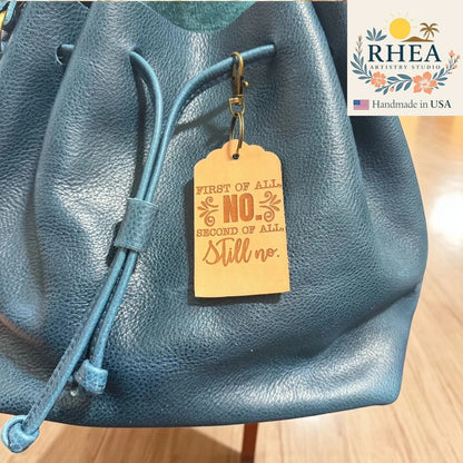 Rhea Artistry Studio natural leather tag engraved with 'First of all, NO. Second of all, Still no.' attached to a teal blue leather bag.