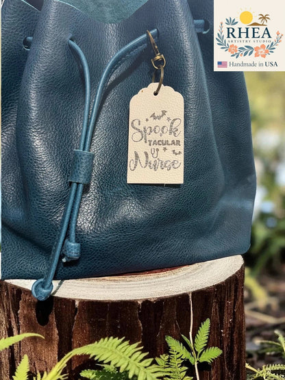 Rhea Artistry Studio cream Saffiano leather tag engraved with 'Spooktacular Nurse' and bats, attached to a teal drawstring bag.
