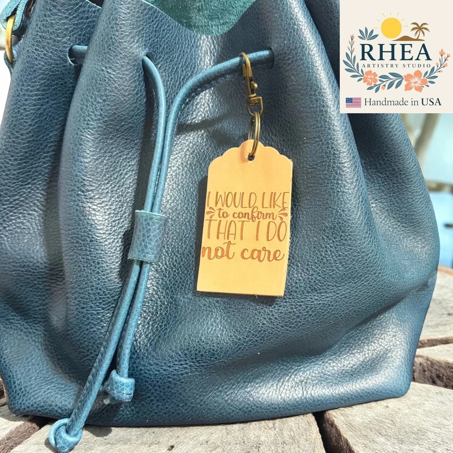 Rhea Artistry Studio tan leather tag engraved with "I would like to confirm that I do not care" attached to a teal blue leather drawstring bag.