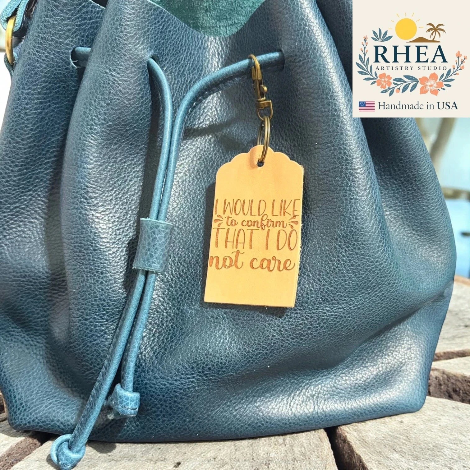 Rhea Artistry Studio tan leather tag engraved with "I would like to confirm that I do not care" attached to a teal blue leather drawstring bag.
