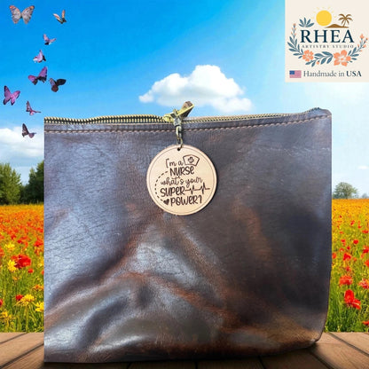 Rhea Artistry Studio light brown leather tag engraved with "I'm a NURSE what's your SUPER POWER?" attached to a dark brown leather bag.