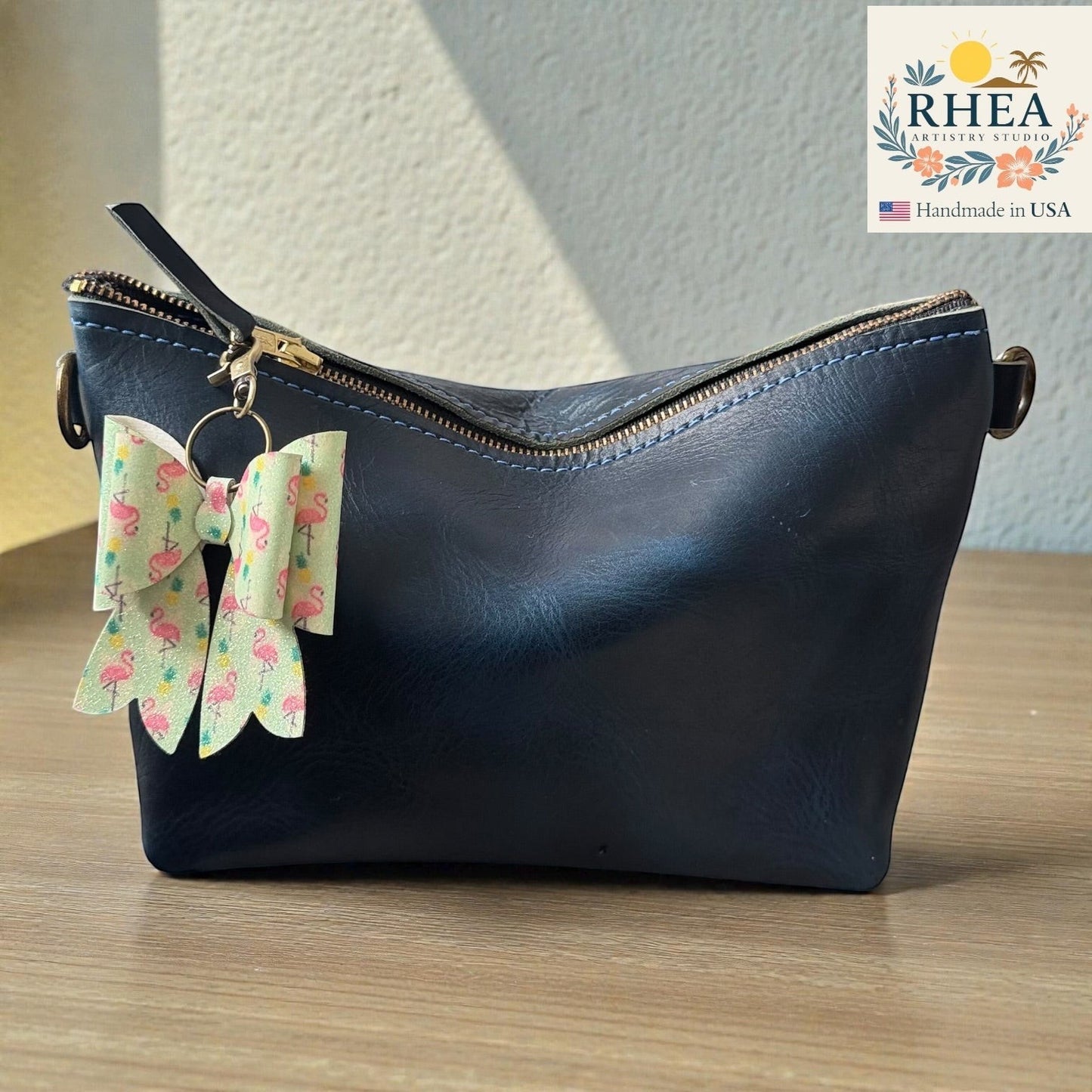 Rhea Artistry Studio handmade double-layer French bow charm with flamingo print and brass hardware, clipped to a navy blue leather bag.