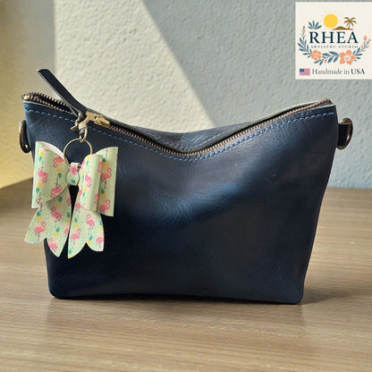 Rhea Artistry Studio handmade double-layer French bow charm with flamingo print and brass hardware, clipped to a navy blue leather bag.
