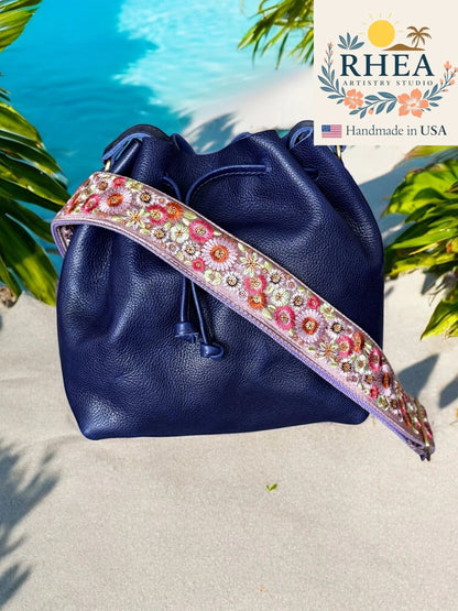 Rhea Artistry Studio navy blue leather bucket bag with a 2-inch wide adjustable floral embroidered strap in pink, purple, and green.
