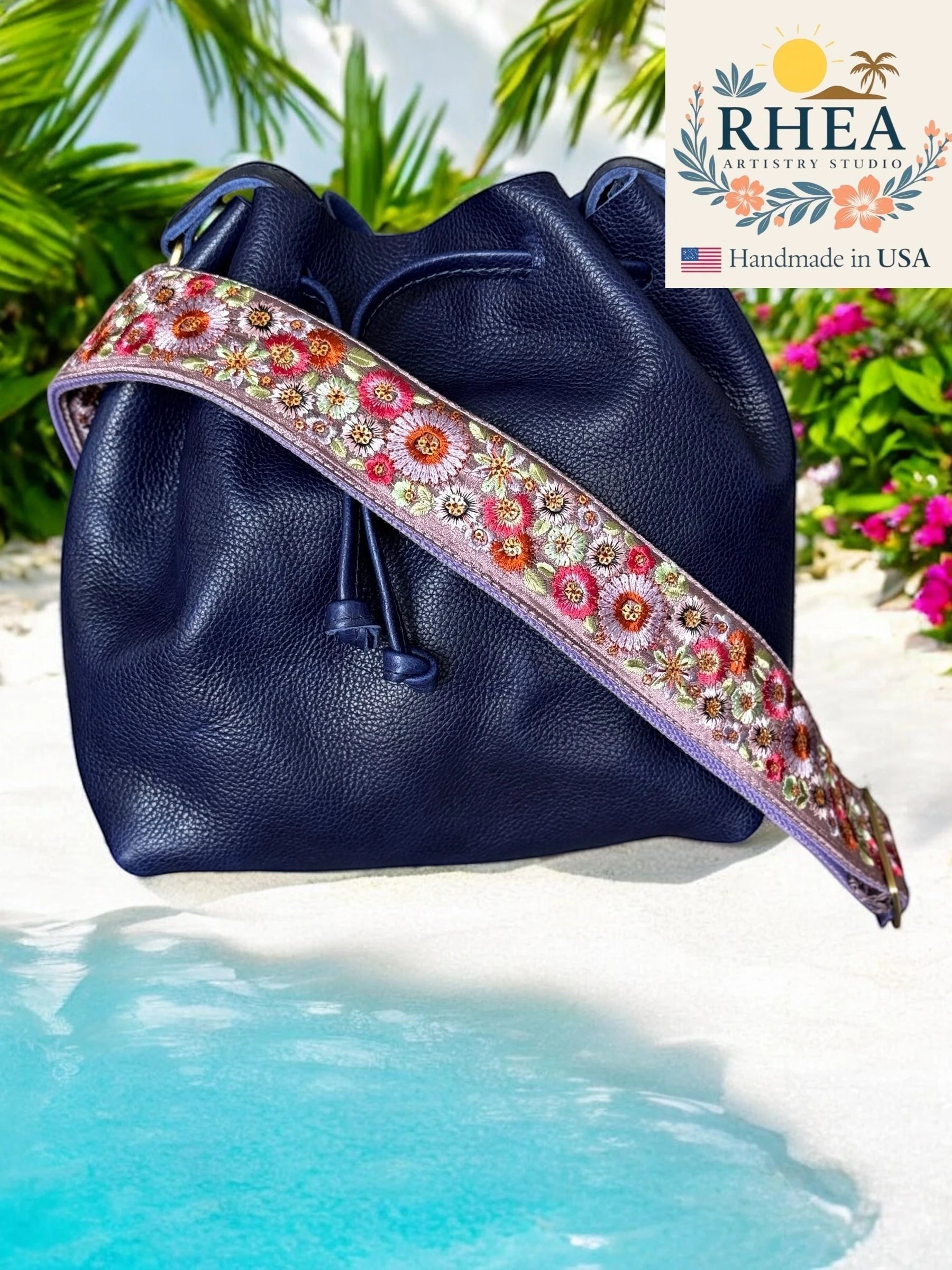 Rhea Artistry Studio navy blue leather bucket bag with a 2-inch wide adjustable floral embroidered strap in pink, white, and orange.