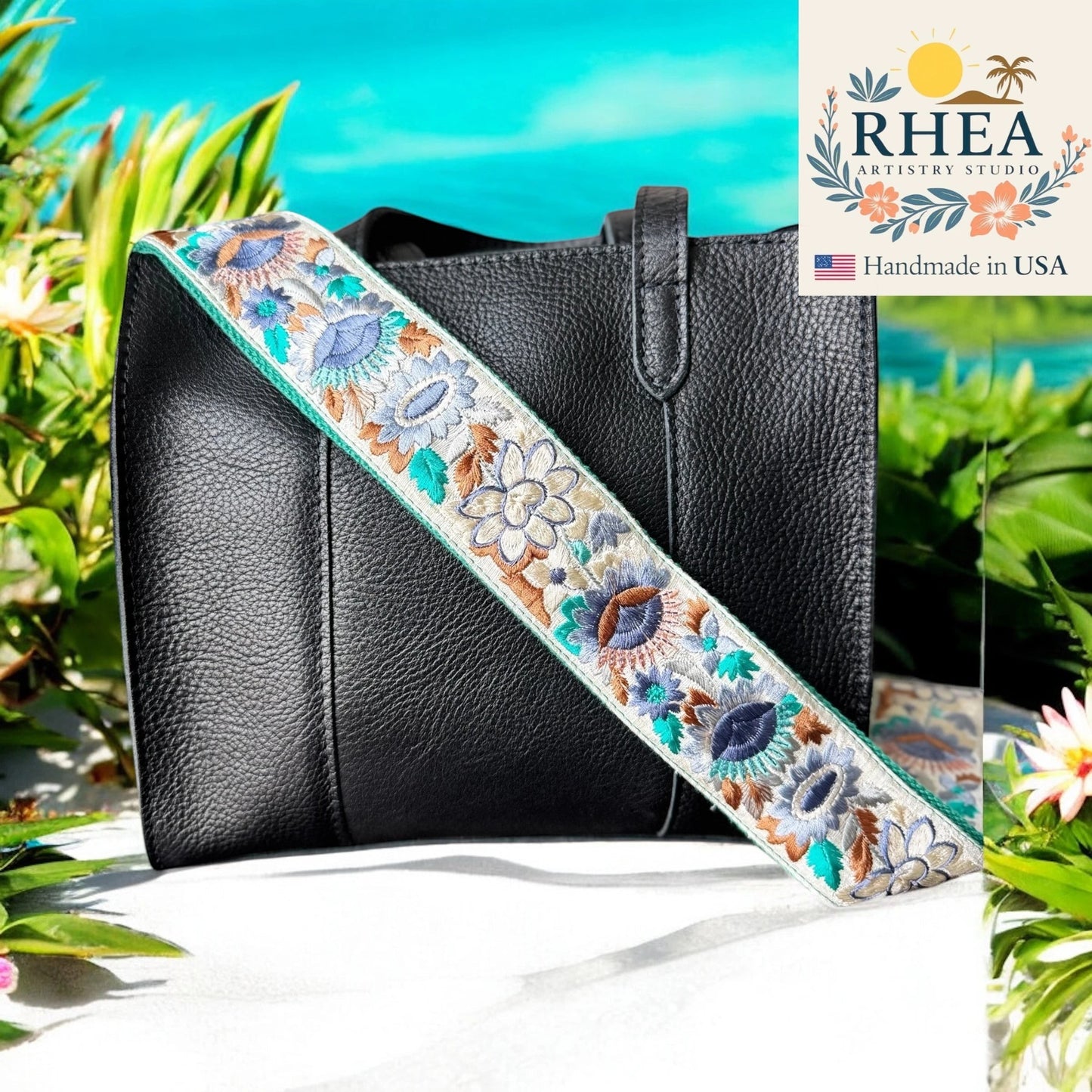Rhea Artistry Studio 2-inch wide crossbody strap with blue and brown floral embroidery on a cream background, adjustable length, brushed brass hardware.