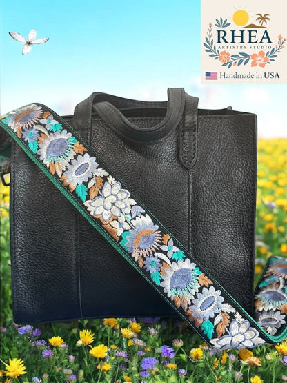 Rhea Artistry Studio 2-inch wide crossbody strap with blue, grey, and teal floral embroidery on black backing, adjustable 30-54 inches, brushed brass hardware.