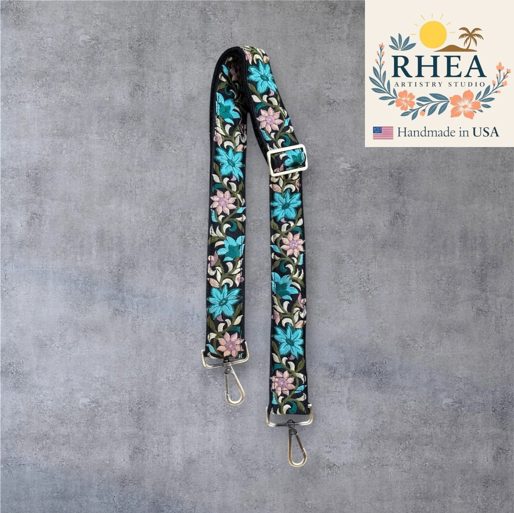 Rhea Artistry Studio black crossbody strap with bold floral embroidery in teal, pink, and green. 1.5-inch width, adjustable length, brushed brass hardware.