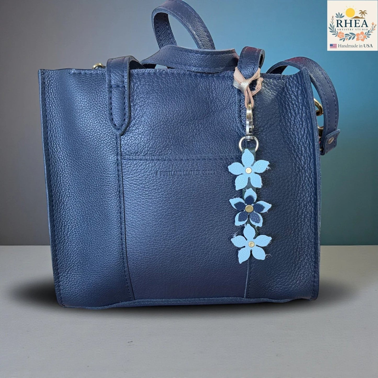 Rhea Artistry Studio navy blue leather tote bag with a handmade floral charm featuring light blue and navy leather flowers and a brushed brass clasp.