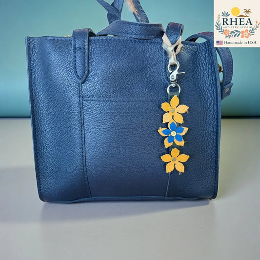Rhea Artistry Studio navy blue leather tote bag with a handmade floral charm featuring yellow and blue leather flowers and a brushed brass clasp.