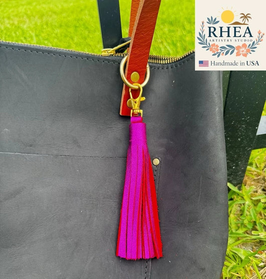 Rhea Artistry Studio large metallic fuchsia leather tassel keychain with gold lobster clasp attached to a black leather tote bag.