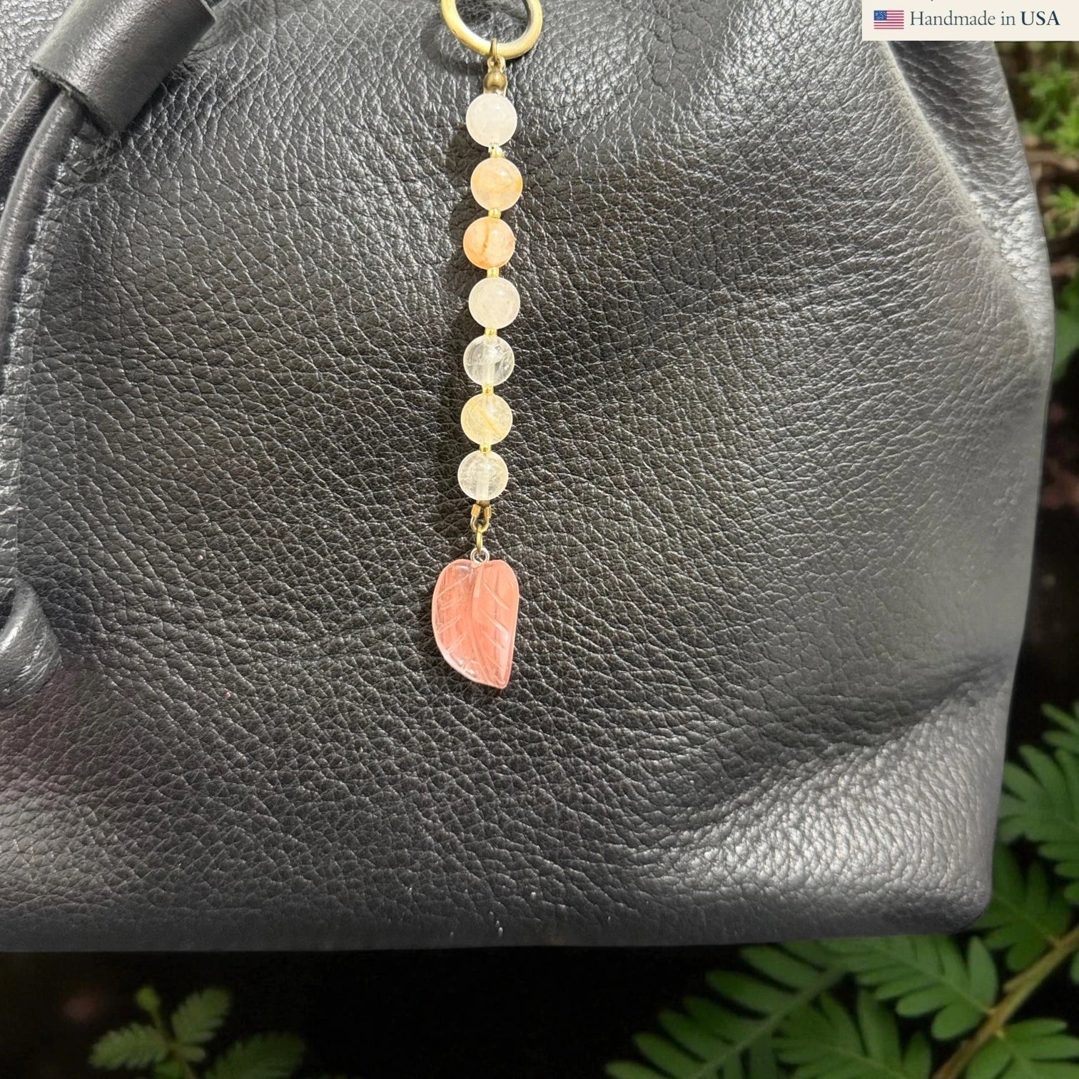 Rhea Artistry Studio handmade gemstone and glass leaf charm with peach and clear beads on a black leather bag.