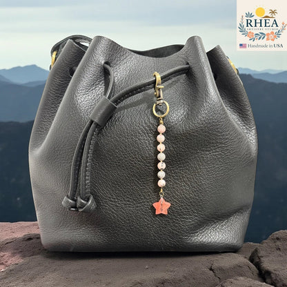 Rhea Artistry Studio handmade bag charm with pink gemstone beads and a coral star pendant, attached to a black leather bucket bag.