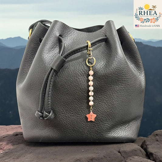 Rhea Artistry Studio handmade bag charm with pink gemstone beads and a coral star pendant, attached to a black leather bucket bag.