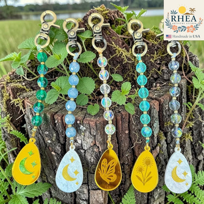 Rhea Artistry Studio handcrafted bag charms with multi-faceted glass beads in green, blue, and clear AB finish, and engraved pendants featuring moons, stars, butterflies, and flowers. Brass hardware.