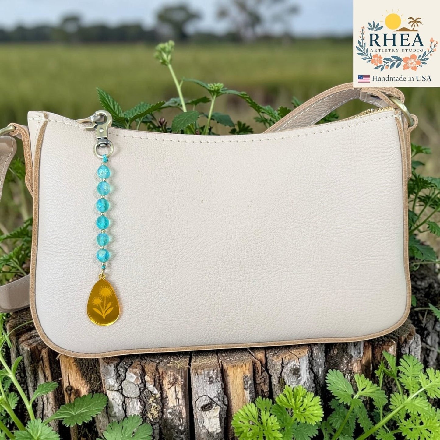Rhea Artistry Studio bag charm with blue faceted glass beads and a gold engraved pendant, attached to a cream leather handbag.