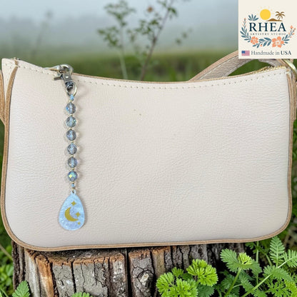 Rhea Artistry Studio bag charm with multi-faceted glass beads and a moon and star pendant, clipped to a cream leather handbag.