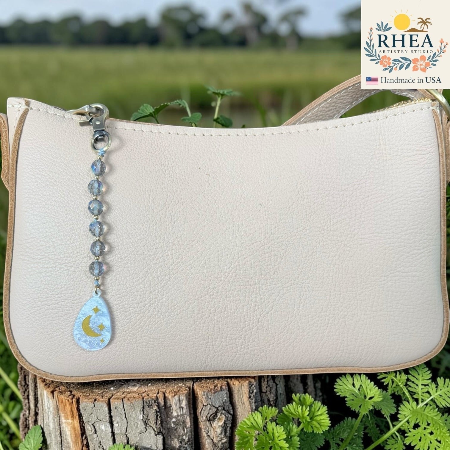 Rhea Artistry Studio bag charm with multi-faceted glass rounds and moon and star pendant, brass hardware, clipped to a cream leather handbag.