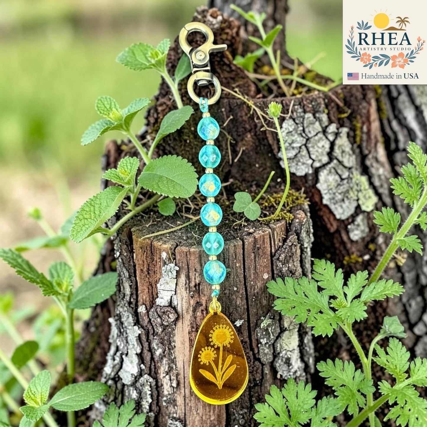 Rhea Artistry Studio handcrafted bag charm with blue multi-faceted glass beads and a yellow teardrop pendant with engraved sunflowers.